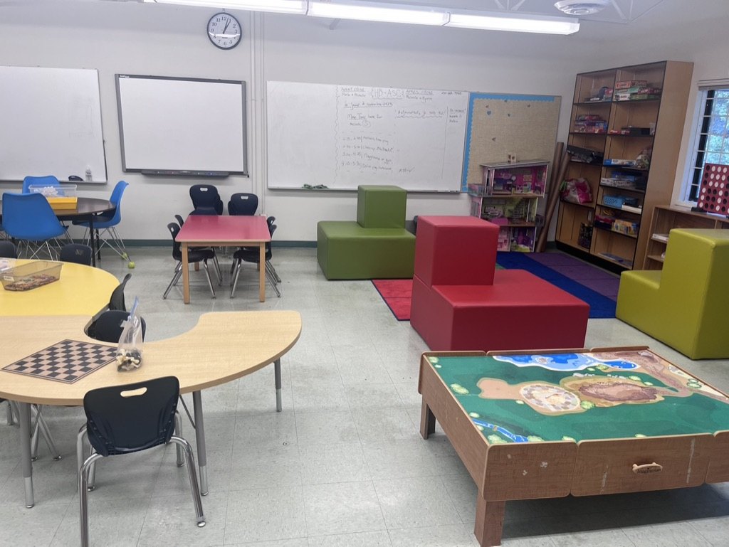 A bright classroom arranged with varied tables, soft seating, games, books, and open floor space